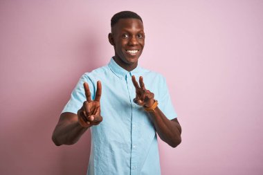 Mavi gömlekli Afro-Amerikan bir adam izole edilmiş pembe arka planda durup kameraya bakıp zafer işareti yaparken gülümsüyor. İki numara..