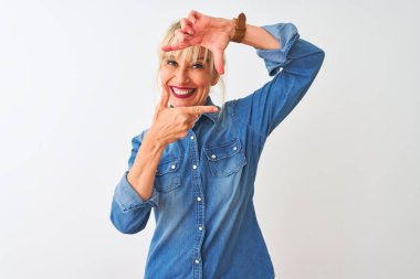 İzole edilmiş beyaz arka planda dikilen sıradan bir kot gömlek giyen orta yaşlı bir kadın gülümsüyor ve elleri ve parmakları mutlu yüzlü bir çerçeve yapıyor. Yaratıcılık ve fotoğrafçılık kavramı.