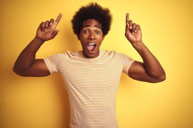 Afro saçlı, çizgili tişörtlü Amerikalı adam izole edilmiş sarı arka planda duruyor. Şaşırmış, şaşırmış ve parmaklarıyla işaret ediyor ve kollarını kaldırıyor..
