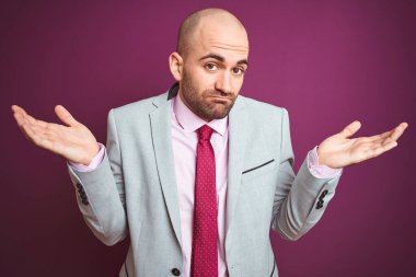 Takım elbiseli ve kravatlı genç bir iş adamı. Mor, soyutlanmış bir arka plan. Kolları ve elleri havada, kafası karışmış bir ifade. Şüphe kavramı.