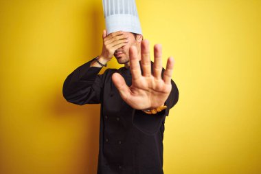 Üniformalı ve şapkalı genç aşçı, izole edilmiş sarı arka planda duruyor, gözleri ellerle örtüyor ve üzgün ve korku ifadeleriyle jestleri durduruyor. Utanç verici ve olumsuz bir kavram..