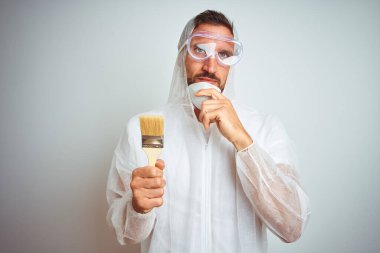 Profesyonel işçi teçhizatı giyen ressam izole edilmiş arka planda fırça tutuyor. Ciddi bir yüz ifadesi var. Soru sormayı düşünüyor.