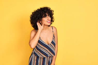 Kıvırcık saçlı, çizgili renkli elbiseler giyen, izole edilmiş sarı arka planda sırıtan, dedikodu ve dedikoduları dinleyen Arap bir kadın. Sağırlık kavramı.