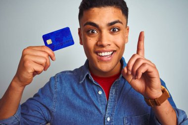 Genç Brezilyalı müşteri kredi kartı tutuyor. İzole edilmiş beyaz arka planda duruyor. Bir fikir ya da soru işareti ile şaşırıyor.