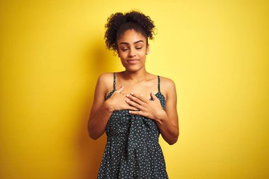 Afrika kökenli Amerikalı bir kadın, izole edilmiş sarı arka plan üzerine günlük yeşil elbiseler giyip elleri göğsünde, gözleri kapalı ve yüzünde minnet dolu jestlerle gülümsüyor. Sağlık kavramı.