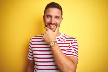 Sarı izole edilmiş arka plan üzerine sıradan kırmızı çizgili tişört giyen genç yakışıklı adam kameraya kollarını kavuşturup gülümseyerek ve çenesini kaldırarak bakıyor. Olumlu düşünüyorum..