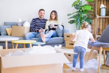 Güzel aile, anne-babalar kanepede kahve içme oturmuş karton kutular etrafında yeni evde oynayan çocuğuna bakıyor