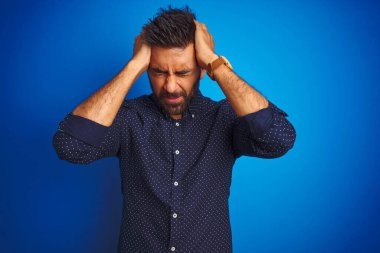Genç Hintli zarif erkek, izole edilmiş mavi arka planda dikilip acı ve migren yüzünden çaresiz ve stresli bir şekilde acı çekiyor. Ellerini başının üstüne koy..