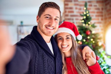Mutlu ve kendinden emin gülümseyen genç bir çift. Evdeki Noel ağacının etrafında kamerayla selfie çekmek.