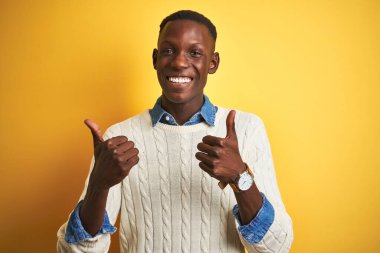İzole edilmiş sarı arka plan işareti üzerine kot gömlek ve beyaz kazak giyen Afro-Amerikalı adam el hareketleriyle olumlu bir hareket yapıyor, gülümsüyor ve mutlu. Neşeli ifade ve kazanan jest.