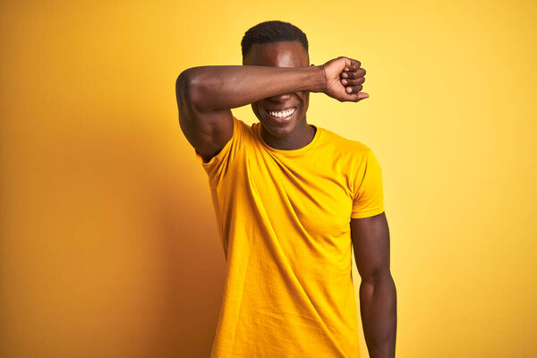 Young african american man wearing casual t-shirt standing over isolated yellow background covering eyes with arm smiling cheerful and funny. Blind concept.