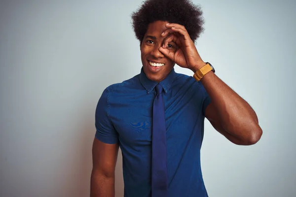 American business man with afro hair wearing blue shirt and tie over isolated white background doing ok gesture with hand smiling, eye looking through fingers with happy face.