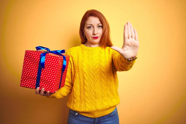 Young beautiful redhead woman holding birthday gift over yellow isolated background with open hand doing stop sign with serious and confident expression, defense gesture