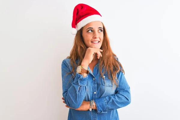 若い美しい赤毛の女性は 集中的な表現について考える顎の手で孤立した背景にクリスマスの帽子をかぶっている 笑顔と思慮深い顔 疑いの概念 — ストック写真