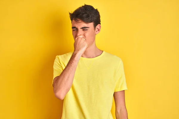 Teenager boy wearing yellow t-shirt over isolated background smelling something stinky and disgusting, intolerable smell, holding breath with fingers on nose. Bad smells concept.
