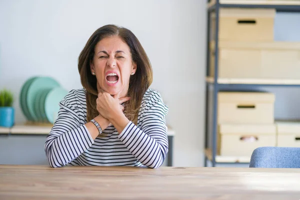 stock image Middle age senior woman sitting at the table at home shouting suffocate because painful strangle. Health problem. Asphyxiate and suicide concept.