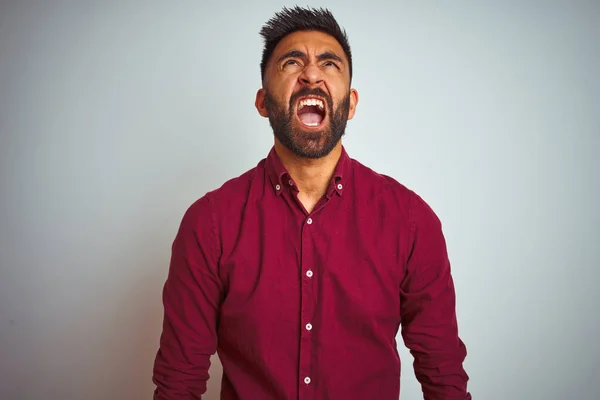 Young indian man wearing red elegant shirt standing over isolated grey background angry and mad screaming frustrated and furious, shouting with anger. Rage and aggressive concept.