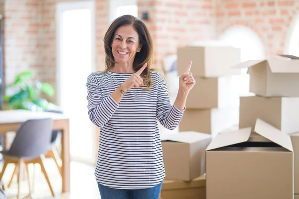Middle age woman moving to a new house arround cardboard boxes smiling and looking at the camera pointing with two hands and fingers to the side.