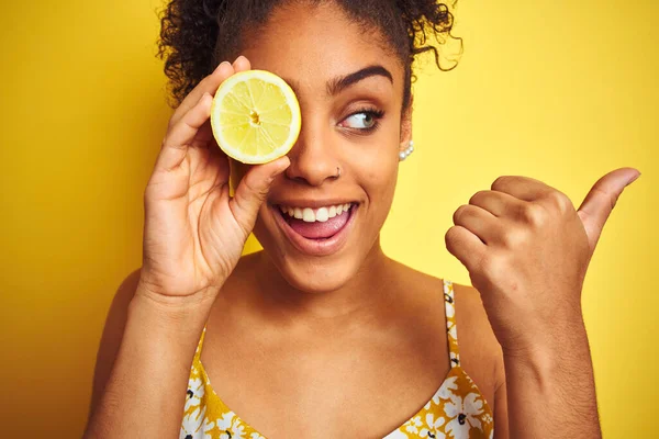 若いアフリカ系アメリカ人の女性がレモンのスライスを持っています孤立した黄色の背景を指し 幸せな顔を笑顔で親指で側面まで示す — ストック写真