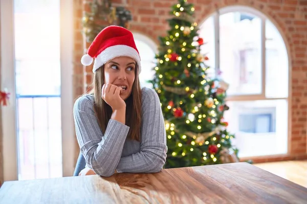 年轻而漂亮的女人 戴着圣爪帽坐在家里的桌子旁 在圣诞装饰前显得紧张而紧张 双手放在咬着钉子的嘴上 焦虑问题 — 图库照片