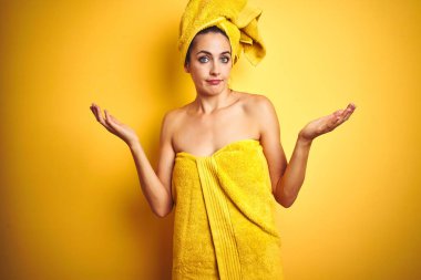 Vücuduna ve başına duş havlusu takmış güzel bir kadın sarı izole edilmiş arka planda bilgisiz ve elleri ve kolları havada kafası karışmış bir ifade. Şüphe kavramı.