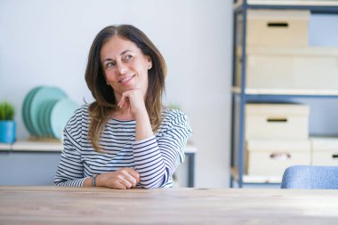 Orta yaş yaşlı kadın evde masada otururken elini çeneüzerinde sorgular, dalgın ifade eder. Gülümseyen ve düşünceli bir yüz. Şüphe kavramı.