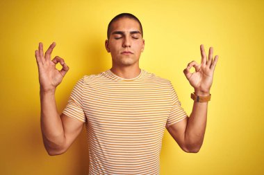 Sarı izole edilmiş arka plan üzerine çizgili tişört giyen yakışıklı genç adam rahatla ve parmaklarıyla meditasyon hareketi yaparken gözleri kapalı gülümse. Yoga konsepti.
