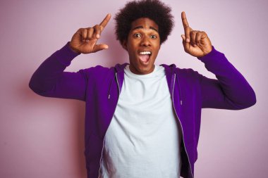 Genç Afro-Amerikalı adam mor kazak giyip pembe arka planda dikilmiş şaşkın şaşkın şaşkın gülümsüyor, parmaklarıyla işaret ediyor ve kollarını kaldırıyordu..
