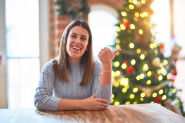 Genç ve güzel bir kadın Noel ağacı ve süsleme masasında oturmuş gülümsüyor ve başparmağıyla yan tarafa bakıyor..
