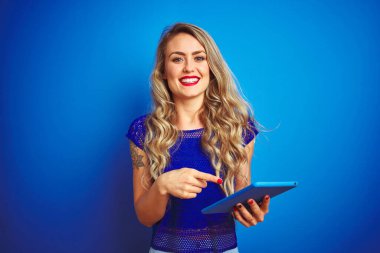 Genç ve güzel bir kadın mavi arka plan üzerinde tablet kullanıyor. Elle ve parmakla işaret ederek çok mutlu.