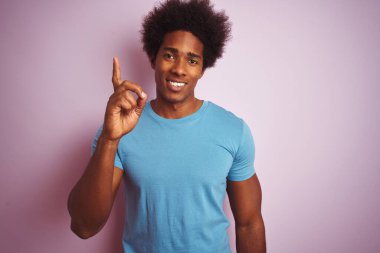 Afro saçlı, mavi tişörtlü, izole edilmiş pembe arka planda duran ve bir numarayı işaret eden Afro-Amerikan adam kendinden emin ve mutlu bir şekilde gülümsüyor..