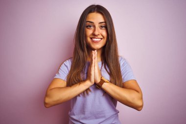 Genç ve güzel bir kadın, günlük tişört giyip pembe arka planda dikilip ellerini birleştirip af diliyor kendinden emin bir şekilde gülümsüyor..