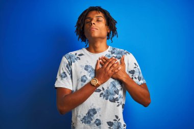 Rasta desenli tişörtlü Afro-Amerikan bir adam izole edilmiş mavi arka plan üzerinde elleri göğsünde, gözleri kapalı ve yüzünde minnet dolu bir jest ile gülümsüyor. Sağlık kavramı.