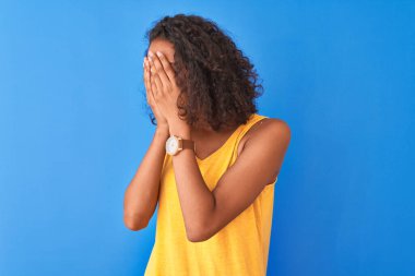 Sarı tişört giyen genç Brezilyalı bir kadın izole edilmiş mavi arka planda dikiliyor. Üzücü yüz ifadesini ellerle kapatıyor ve ağlıyor. Depresyon kavramı.