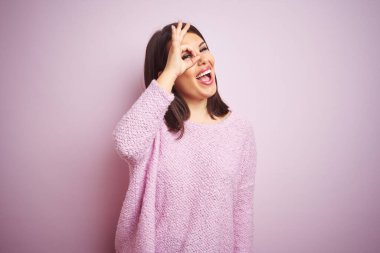Genç, güzel, esmer bir kadın pembe bir arka plan üzerine kazak giyiyor. El hareketleriyle gülümsüyor ve mutlu yüzüyle parmaklarının arasından bakıyor..