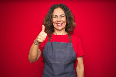 Kırmızı izole arka planda önlük giyen orta yaşlı bir kadın mutlu bir el hareketi yapıyor. Kameraya bakıp başarıyı gösteren ifade onaylanıyor.
