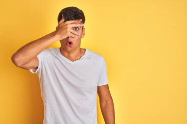 İzole edilmiş sarı arka plan üzerinde duran beyaz t-shirt giyen genç Hintli adam şok içinde yüzünü ve gözlerini kapayarak utangaç ifadesiyle parmaklarının arasından bakıyor..