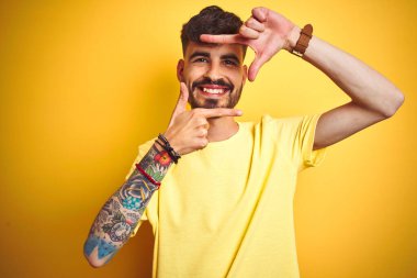 İzole edilmiş sarı arka planda duran tişörtlü genç adam gülümsüyor ve mutlu yüzlü elleri ve parmakları olan bir çerçeve yapıyor. Yaratıcılık ve fotoğrafçılık kavramı.