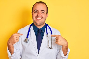 Paltolu ve steteskoplu genç bir doktor izole edilmiş sarı arka planda duruyor. Yüzünde bir gülümsemeyle, gururlu ve mutlu bir şekilde kendini işaret ediyor..