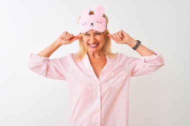 İzole edilmiş beyaz arka plan üzerinde uyku maskesi ve pijama giyen orta yaşlı bir kadın yüksek sesli müzik sesi için parmaklarıyla kulaklarını kapatıyor. Sağır kavramı.