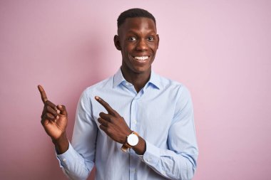 Mavi şık gömlekli Afro-Amerikan bir adam pembe arka planda dikiliyor gülümsüyor ve kameraya bakıyor iki eliyle ve parmaklarıyla yan tarafa bakıyor..