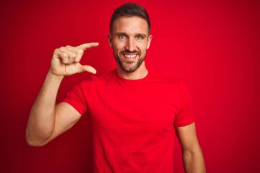 Kırmızı izole edilmiş arka plan üzerine gündelik tişört giyen genç yakışıklı adam gülümsüyor ve el hareketleriyle küçük beden işareti yaparak kameraya bakıyor. Ölçü kavramı.