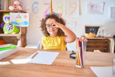 Gözlüklü ve tek boynuzlu at diademi takmış güzel bir çocuk anaokulunda oturmuş el hareketleriyle gülümsüyor ve mutlu yüzüyle parmaklarının arasından bakıyor..