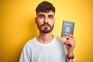 Dövmeli genç bir adam, Amerika Birleşik Devletleri pasaportu ile izole edilmiş sarı arka plan üzerinde, akıllı yüzünde kendine güvenen bir ifadeyle ciddi düşünüyor.