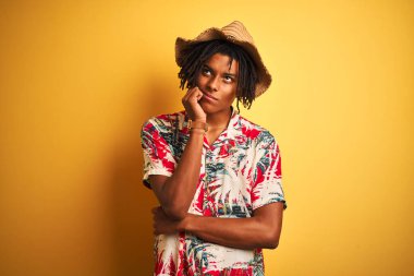 Saçlı, çiçekli tişörtlü ve şapkalı, izole edilmiş sarı arka planı olan, elleri çenesinde soru soran, düşünceli bir Afro-Amerikan adam. Düşünceli bir yüzle gülümsüyor. Şüphe kavramı.