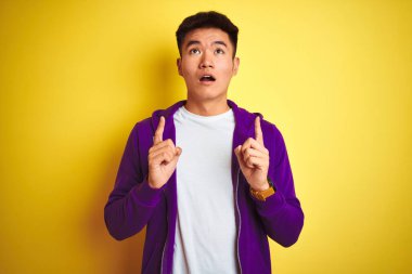 Mor kazak giyen Asyalı genç adam, izole edilmiş sarı arka planda duruyor. Şaşırmış ve şaşırmış bir şekilde yukarı bakıyor ve parmaklarıyla işaret ediyor ve kollarını kaldırıyor..