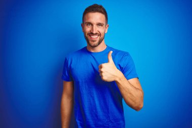 Mavi izole edilmiş arka plan üzerine gündelik tişört giyen genç yakışıklı adam mutlu bir el hareketi yapıyor. Kameraya bakıp başarıyı gösteren ifade onaylanıyor.