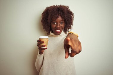 Genç Afrikalı bir kadın elinde kahve fincanıyla izole edilmiş beyaz arka planda dikilip kamerayı işaret ediyor ve sen, el işareti, ön taraftan pozitif ve kendinden emin bir hareket.