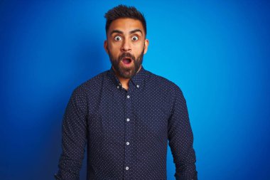Soyutlanmış mavi arka plan üzerinde duran genç Hintli zarif adam korku, korku ve heyecan dolu yüz ifadesiyle korkmuş ve şok olmuştu..