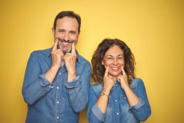 Güzel bir orta yaşlı çift, izole edilmiş sarı arka planda duruyorlar. Ağızları açık gülümsüyorlar, parmakları işaret ediyor ve neşeli bir gülümseme sergiliyorlar.